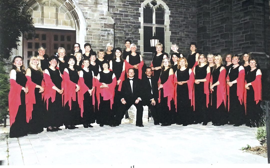 A group photo of the choir in black dresses and red shales outside a grey stone church.