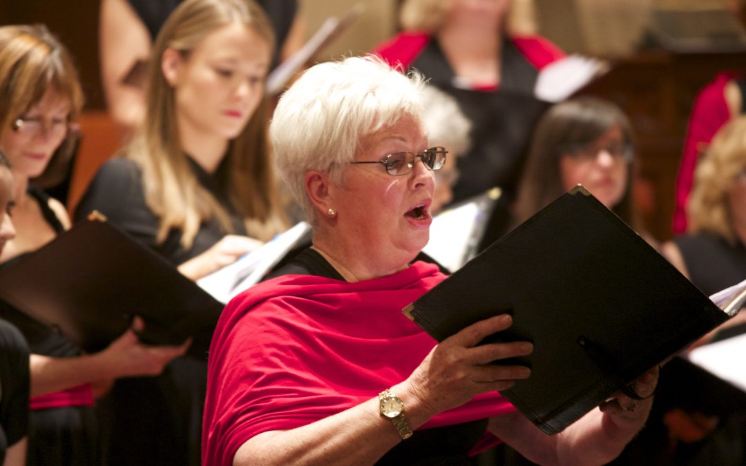 A colour photo of a woman singing a solo and holding a music folder, standing in front of a choir