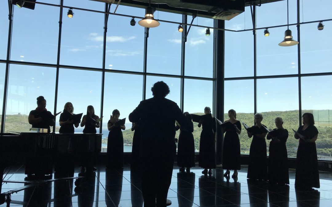 A choir in silhouette performs against a wall of windows overlooking green hills and an ocean inlet.