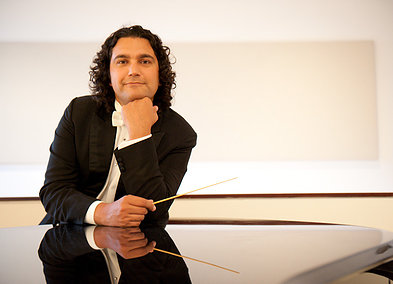 A photo of a man dressed in a tuxedo resting his chin on his left hand, elbow supported on a shiny black grand piano. He is casually holding a conductor's baton in his right hand, resting on the piano.